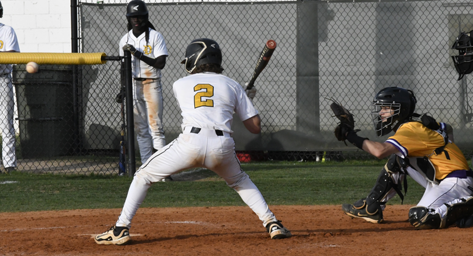 Baseball player at bat in white uniform, catcher in yellow crouched behind, with an approaching pitch and other players in the background near a chain-link fence.