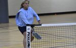 Woman playing pickleball indoors, wearing a light blue long-sleeve shirt and shorts, hitting the ball near the net on a gym floor.