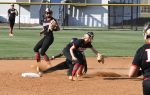 Softball players in action around a base, one sliding into the base as two fielders prepare to catch the ball on a dirt infield.