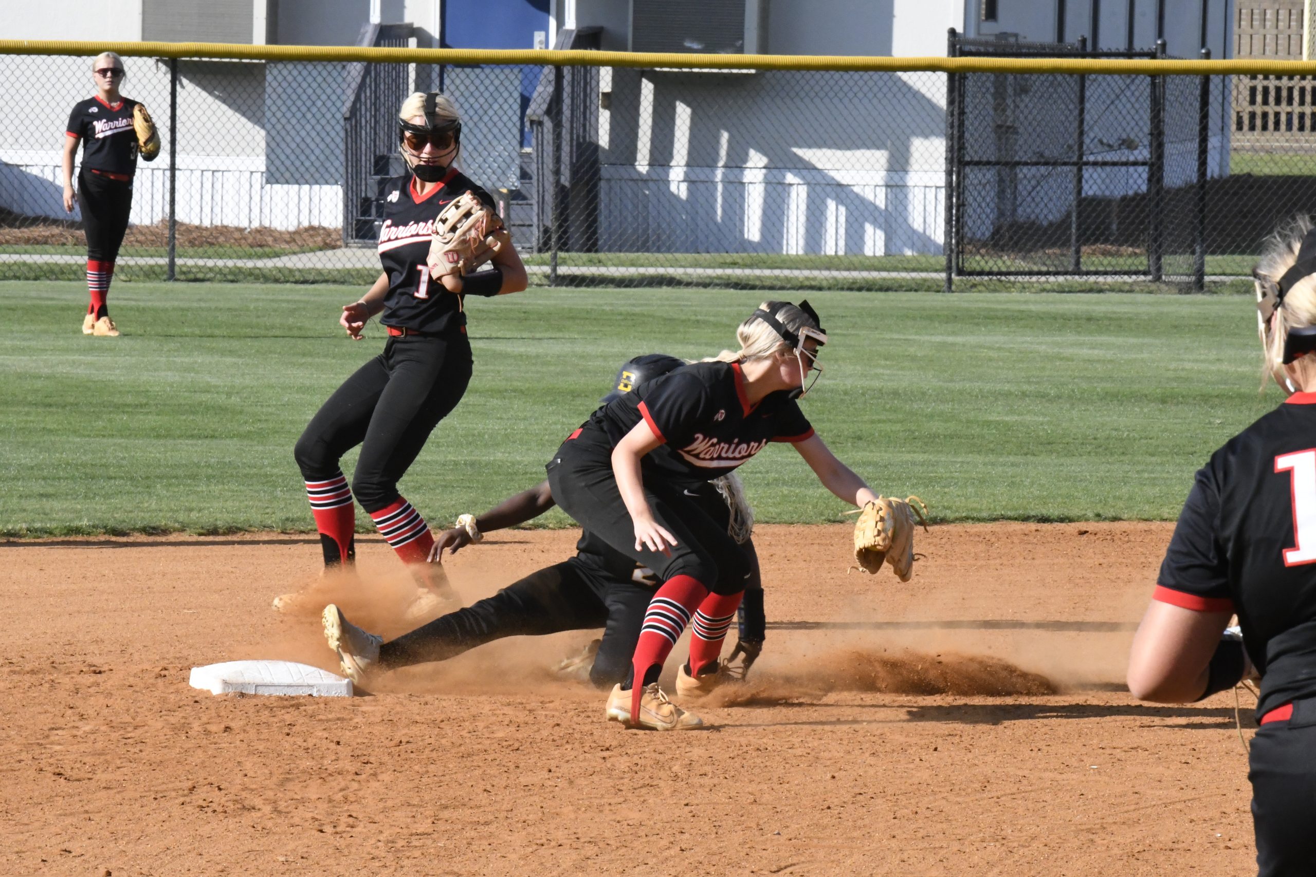 Softball players in action around a base, one sliding into the base as two fielders prepare to catch the ball on a dirt infield.