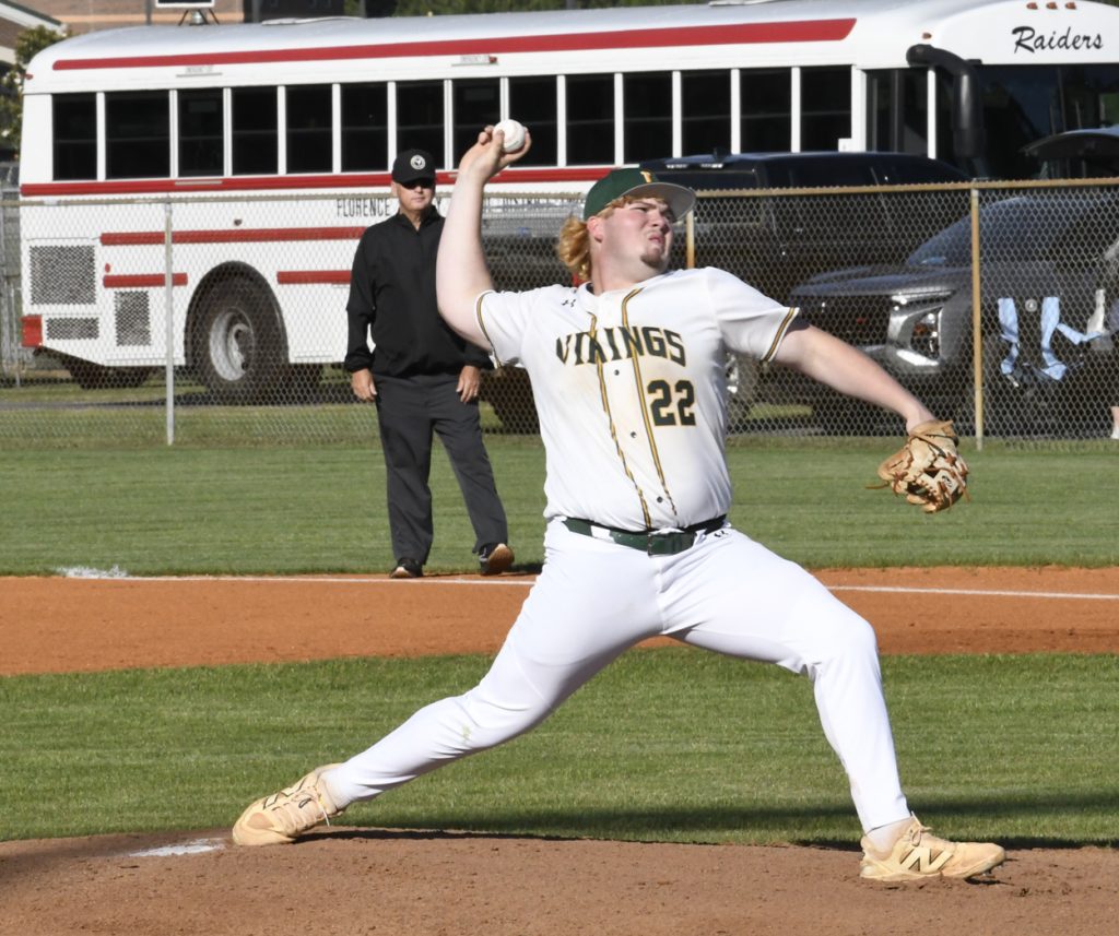 A baseball pitcher in a white "Vikings" uniform throws the ball during a game, with an umpire and school bus in the background.