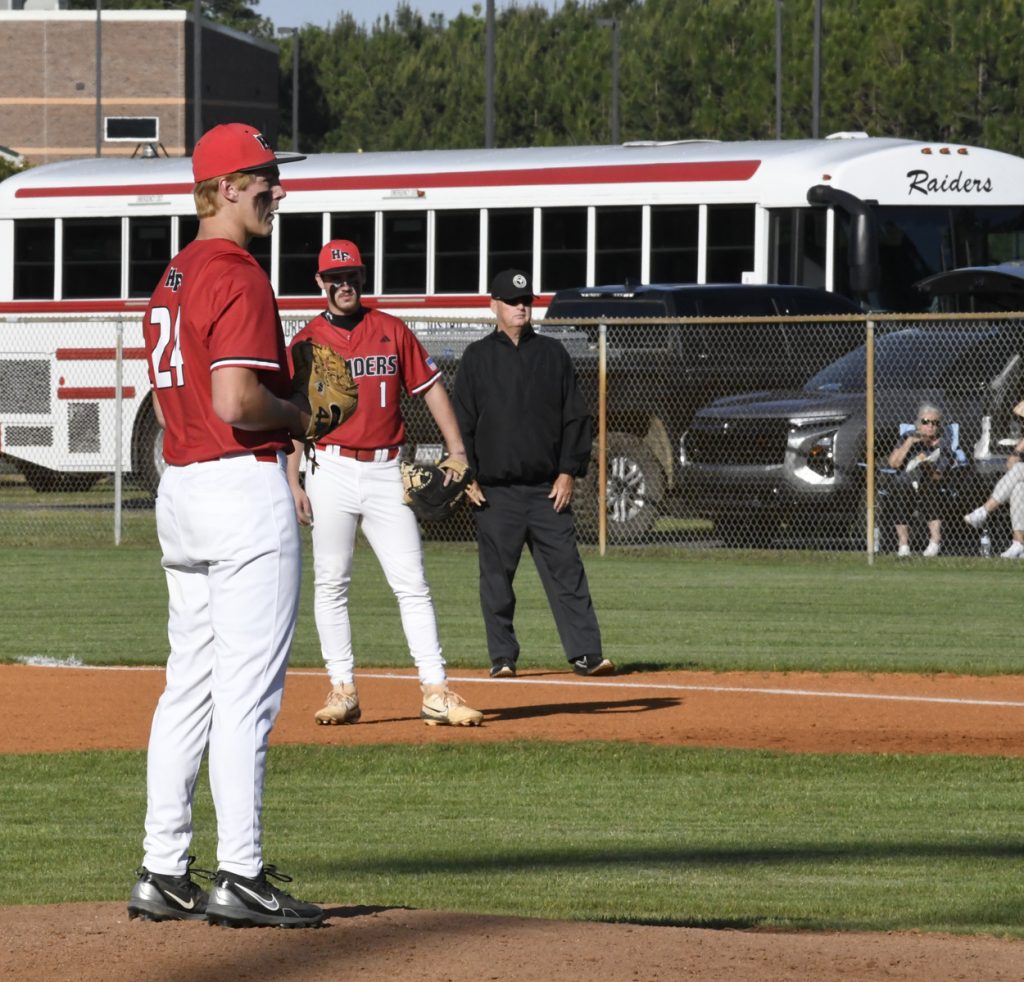 Baseball players in red uniforms and an umpire stand on the field with a school bus and spectators in the background.