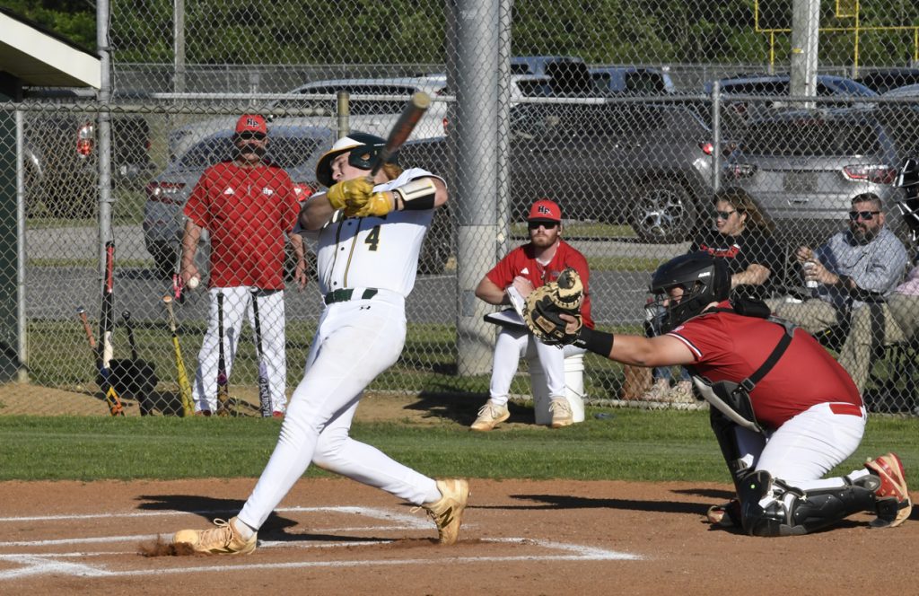 A baseball player in white swings at a pitch as the catcher and umpire watch, with coaches and spectators visible behind a chain-link fence.