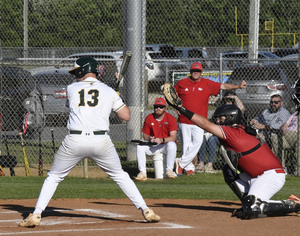 Baseball player at bat with number 13, catcher in red gear, coach and spectators behind fence on a sunny day.