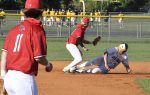 Baseball player in white sliding into base as two players in red uniforms attempt to tag him out, with teams and spectators in the background.