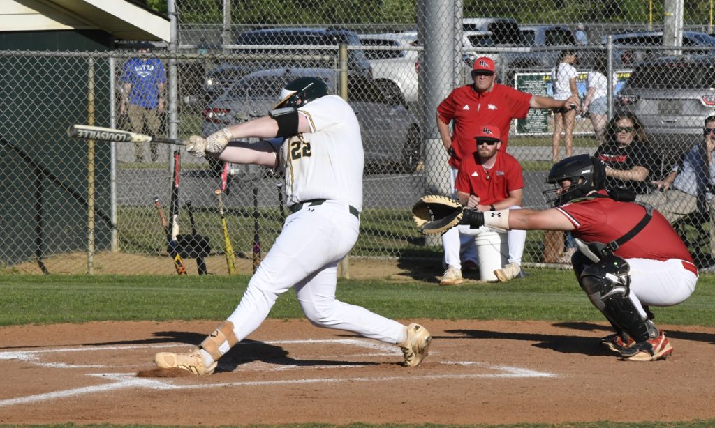 A baseball player in a white uniform swings at a pitch while a catcher and coaches in red watch behind home plate.