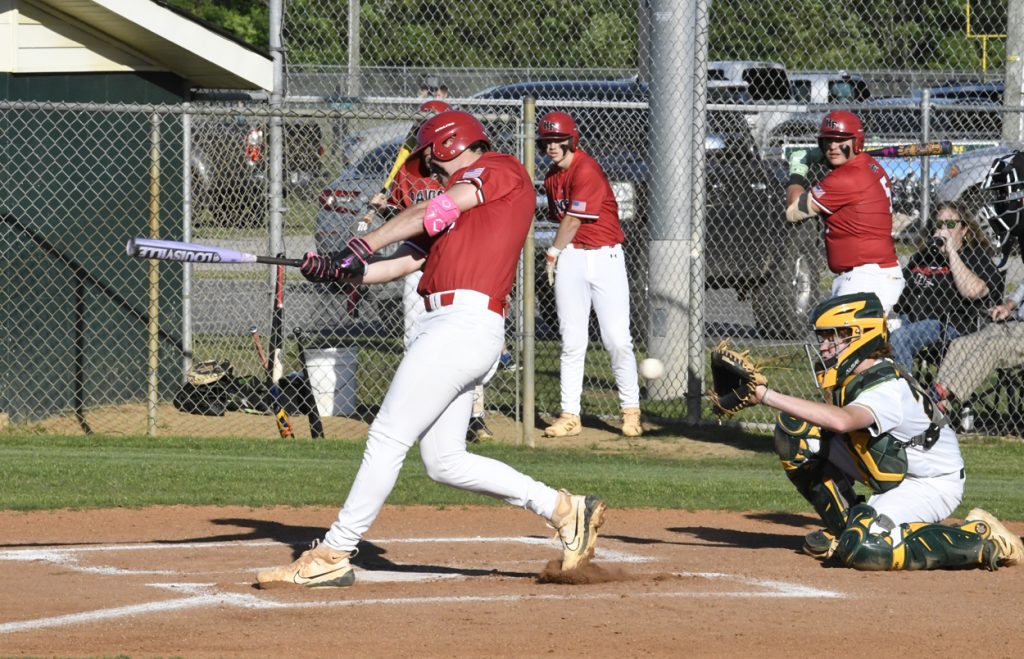 Baseball player in red swinging bat at a pitch, catcher in green gear crouched, teammates watching from the dugout, chain-link fence in background.