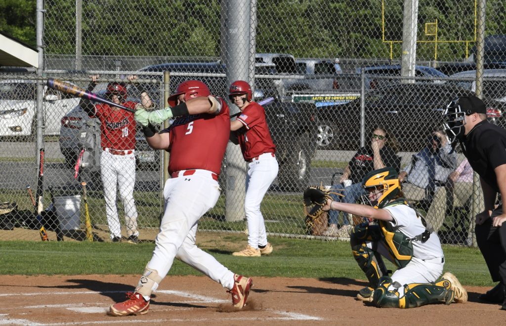 Baseball player in red jersey swings bat at pitch, catcher in green and yellow gear, umpire behind plate, teammates and spectators in background.