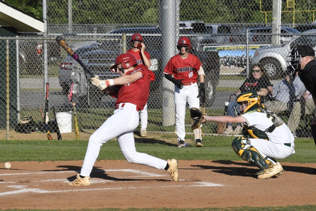 Baseball player in red swinging bat at home plate, catcher in green and yellow gear ready to catch, two teammates and spectators in the background.