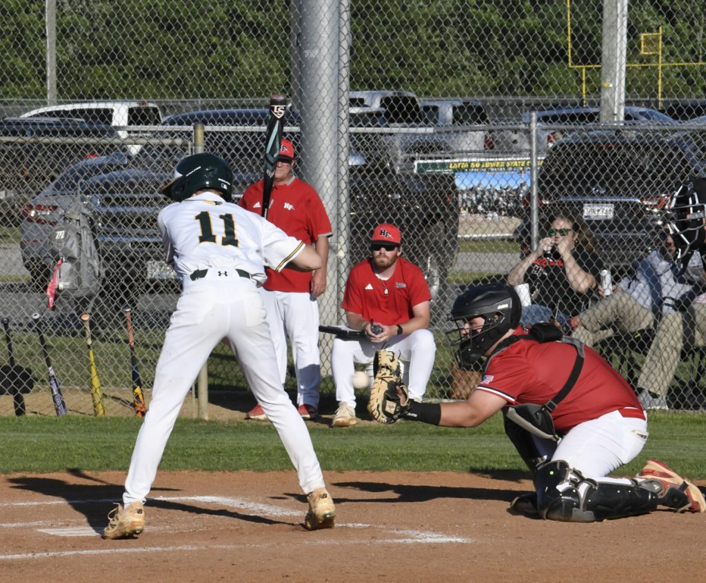 A baseball player in white prepares to bat as a catcher in red crouches, with coaches and spectators watching behind a fence.