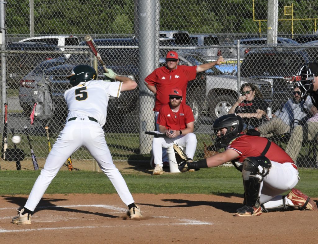 A baseball player in a white uniform prepares to swing at a pitch, while the catcher and umpire are ready behind home plate. Coaches and fans observe.