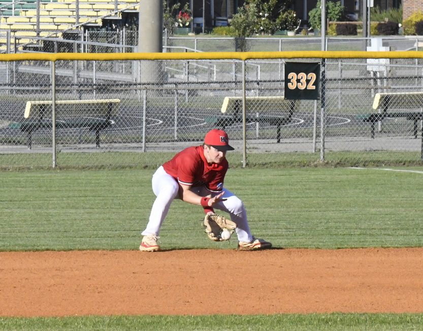 Baseball player in red uniform fielding a ground ball on a sunny baseball field, with empty bleachers and a 332-foot sign in the background.
