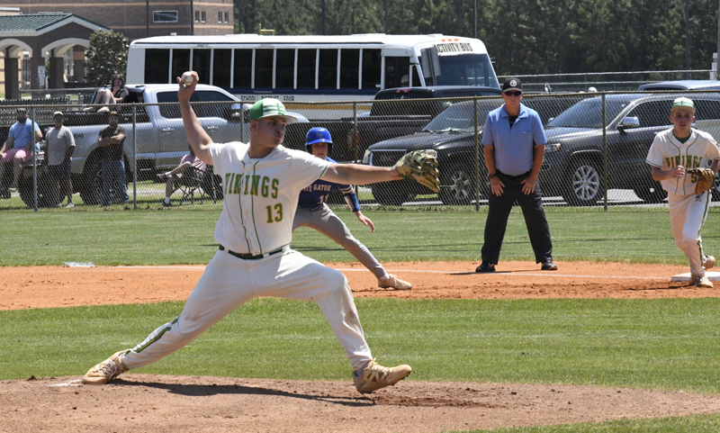 Baseball pitcher in Vikings uniform throws a pitch, while teammates, umpire, and base runner watch. School buses and spectators are in the background.