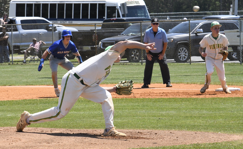 A baseball pitcher throws the ball during a game. Players, an umpire, and a runner are on the field with a school bus and spectators in the background.