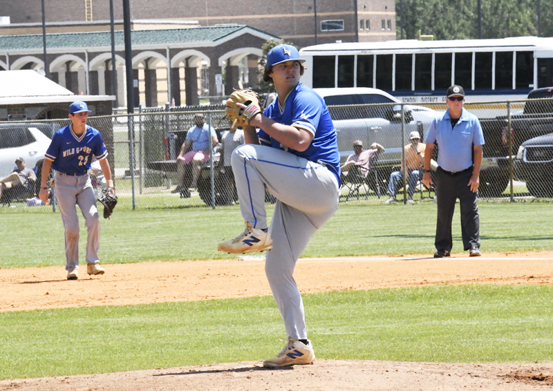 Baseball pitcher in blue uniform winding up to throw, with infielder, umpire, and spectators in background on a sunny day.