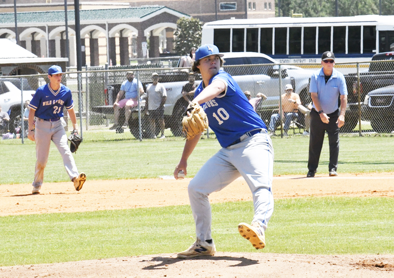 A baseball pitcher in blue uniform throws the ball, with teammates and an umpire on the field. Spectators and a bus are visible in the background.