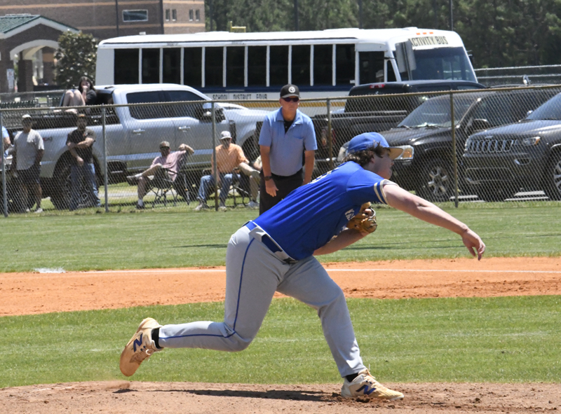 A baseball pitcher in blue uniform throws a pitch on a sunny field, with an umpire, spectators, cars, and a yellow school bus in the background.