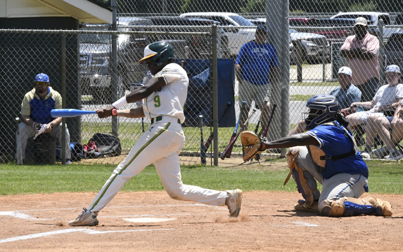 A baseball player in white swings at a pitch while a catcher in blue prepares to catch the ball, with spectators and teammates in the background.