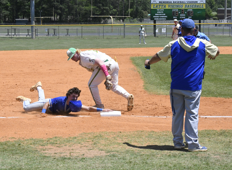 A baseball player in blue slides to third base as a player in white attempts to tag him, with coaches and umpires observing nearby.