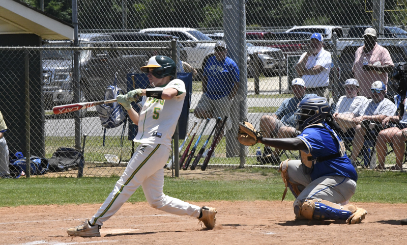 Baseball player in white uniform swinging bat at pitch, catcher in blue gear ready, spectators and parked cars visible behind chain-link fence on a sunny day.