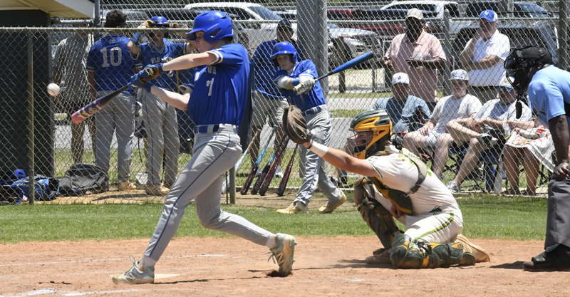 A baseball player in blue swings at a pitch while the catcher in yellow and white crouches behind, with spectators and teammates in the background.