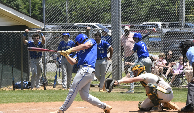A baseball player in blue swings at a pitch while a catcher in green gear crouches behind, with teammates and spectators in the background.
