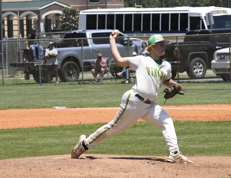 Baseball pitcher in white and green uniform throws a pitch on a sunny day, with spectators and vehicles visible in the background near a fence.