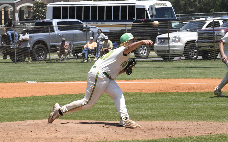 A baseball pitcher in a white and green uniform throws a pitch during a game, with spectators and cars visible in the background.