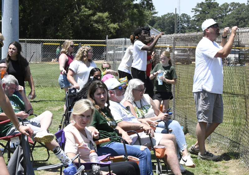 People sitting and standing by a chain-link fence at an outdoor sports event, some watching attentively, others talking, on a sunny day.