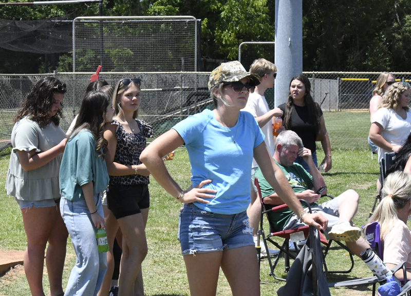 A group of people stand and sit outside near a chain-link fence, watching an event on a sunny day. One woman in a blue shirt stands in the foreground.