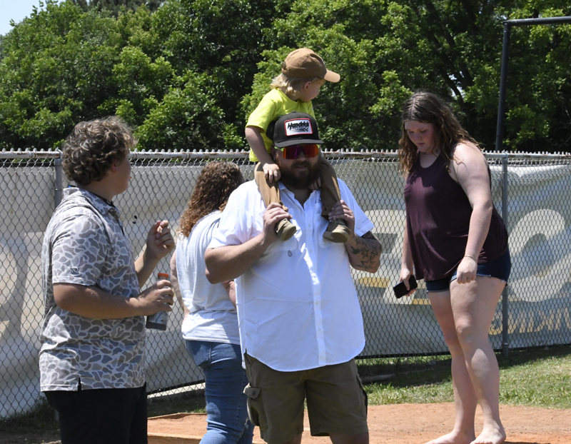 A man carries a child on his shoulders while others stand nearby outside on a sunny day, next to a chain-link fence.