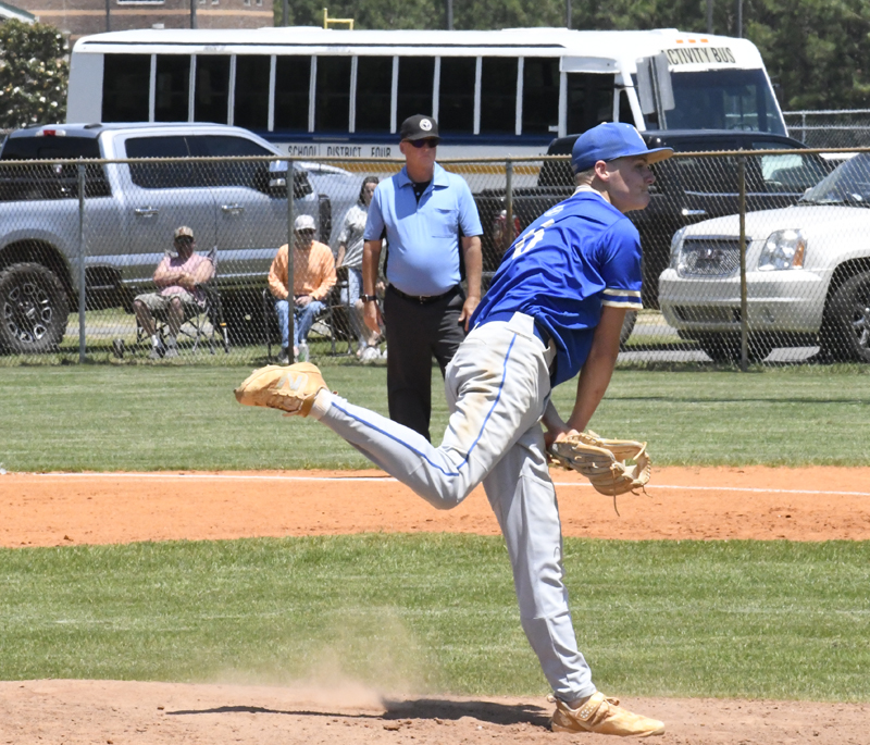Baseball pitcher in blue uniform mid-throw on a sunny field, with umpire, spectators, vehicles, and a school bus in the background.