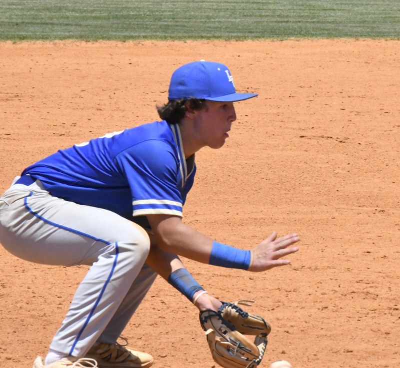 Baseball player in blue uniform crouches on the field, ready to catch a ground ball with glove, during a sunny game.