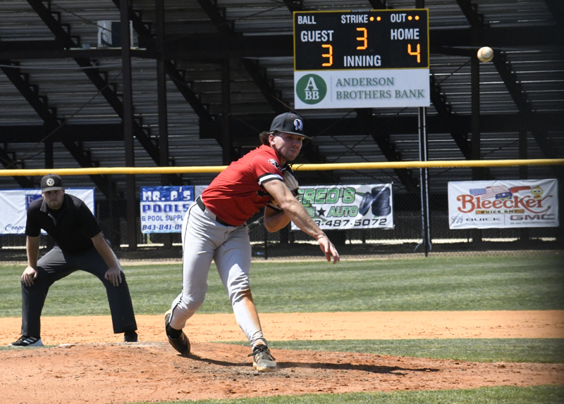 Baseball pitcher in red jersey throws a pitch, umpire watching, scoreboard in background shows home team leading 4-3 in the third inning.
