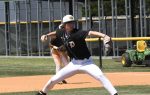 Baseball player in black and white uniform pitching on a field, with another player and sports equipment visible in the background.