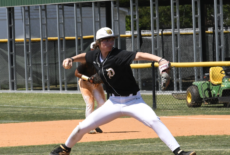 Baseball player in black and white uniform pitching on a field, with another player and sports equipment visible in the background.