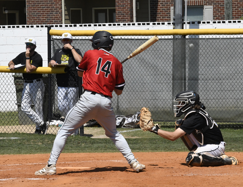 Baseball player in red jersey batting, catcher in black gear behind him, and teammates watching from the dugout, during a daytime game.
