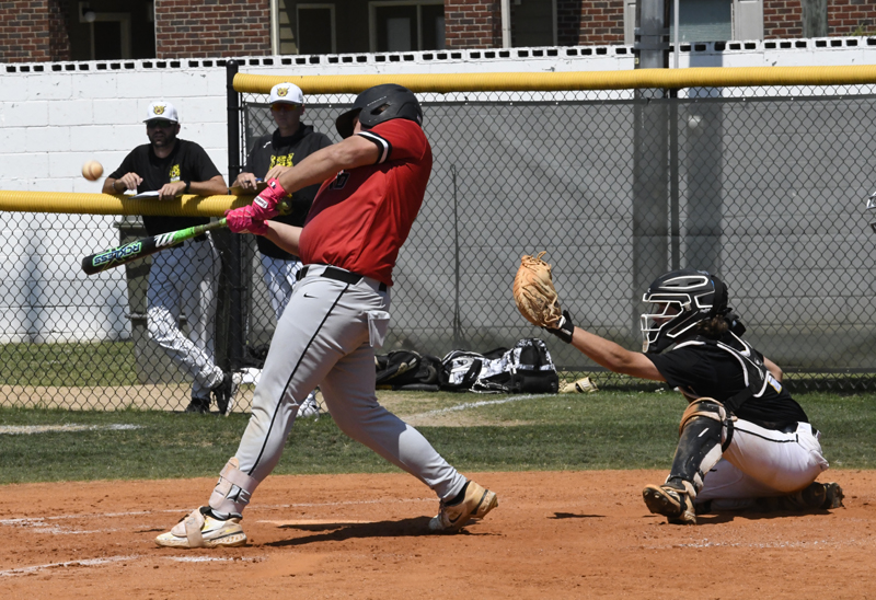 A baseball player in a red jersey swings at a pitch while the catcher and two teammates watch from behind the fence.