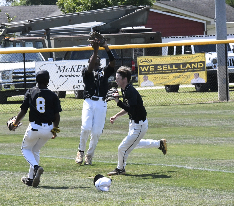 Three youth baseball players in black jerseys converge in the outfield as one jumps to catch a fly ball; a hat lies on the grass nearby.