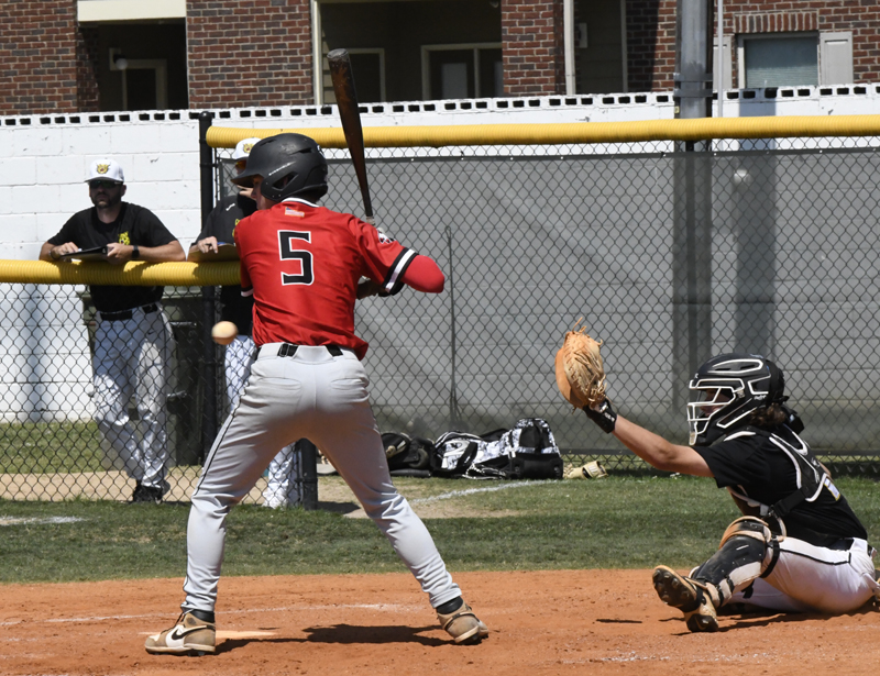 A baseball player in a red jersey prepares to bat as the pitcher throws, with the catcher ready and a coach observing from behind the fence.