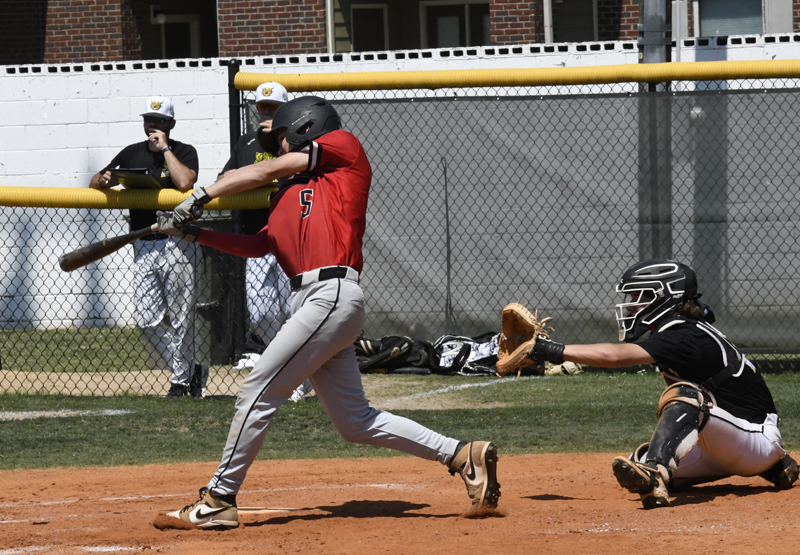 Baseball player in red jersey swinging bat at pitch, catcher in black gear ready to catch, two people watching from behind the fence.