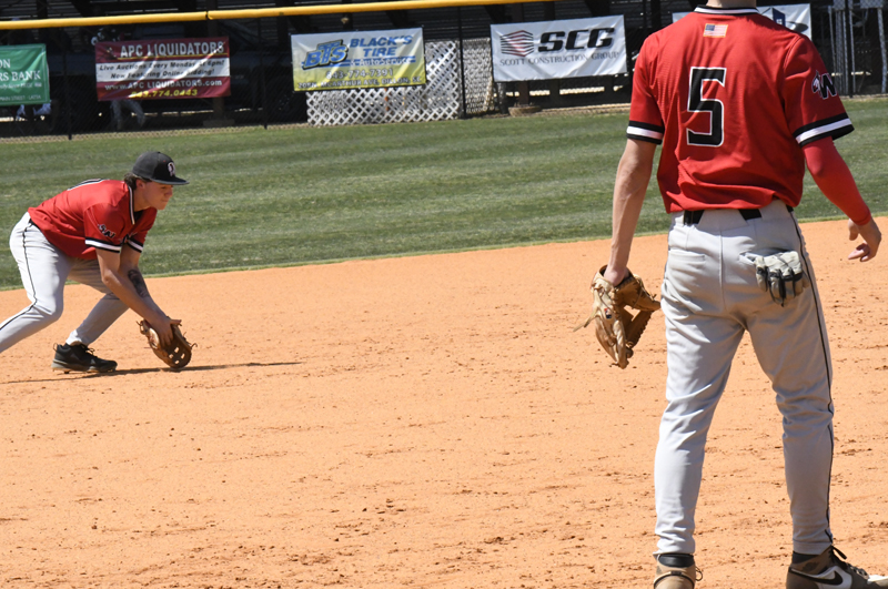 Two baseball players in red jerseys on a field, one preparing to field a ground ball, the other standing nearby with a glove.