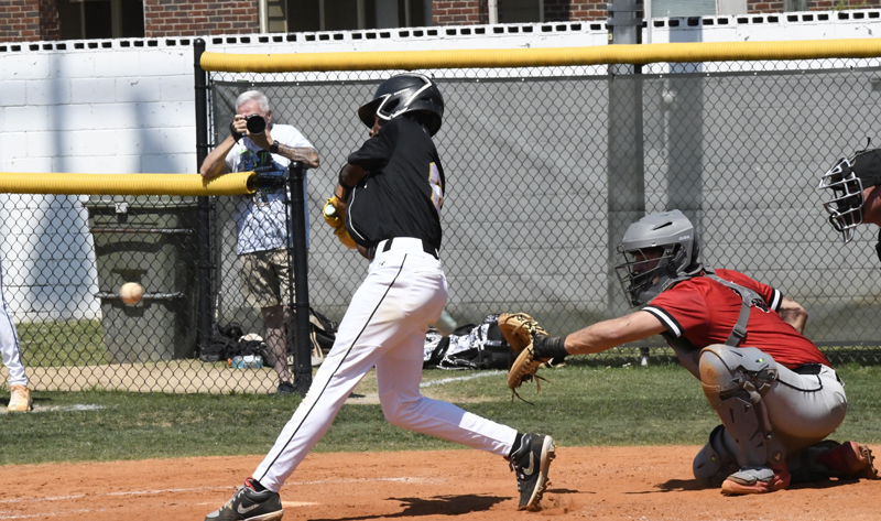 A baseball player in black swings at a pitch, with a catcher in red crouched behind and a photographer capturing the action through a fence.