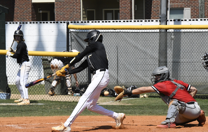 Baseball player in black uniform swings at pitch while catcher in red gear crouches behind home plate, with batter on deck and teammates in background.