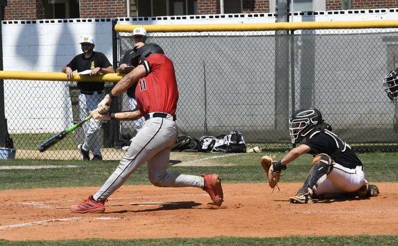 Baseball player in red jersey swinging bat at pitch, catcher crouched behind plate, two people watching from outside fence in sunny outdoor field.