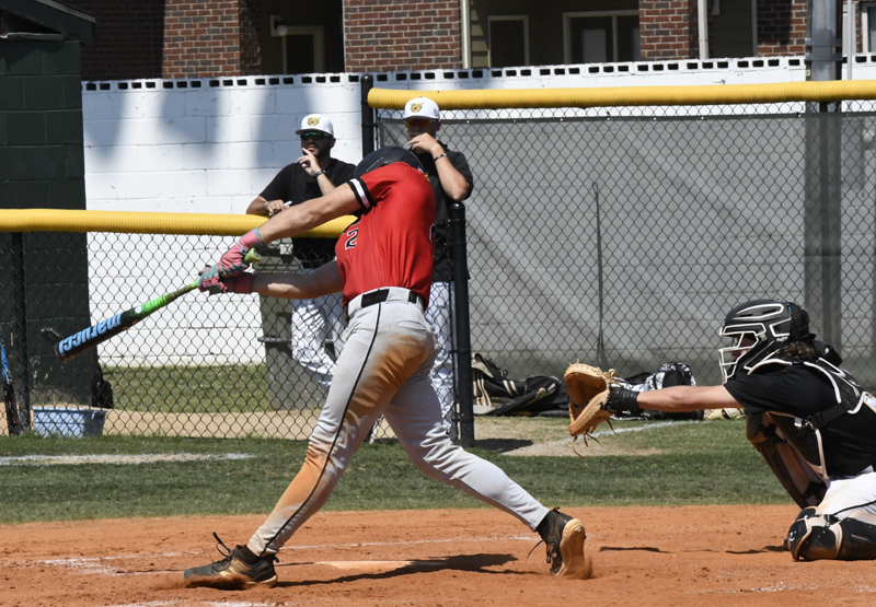 Baseball player in red jersey swings bat at pitched ball, catcher in black gear prepares to catch, two teammates watch behind chain-link fence on sunny day.