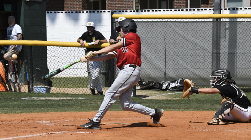 A baseball player in a red jersey swings at a pitch while the catcher and umpire watch closely behind home plate.