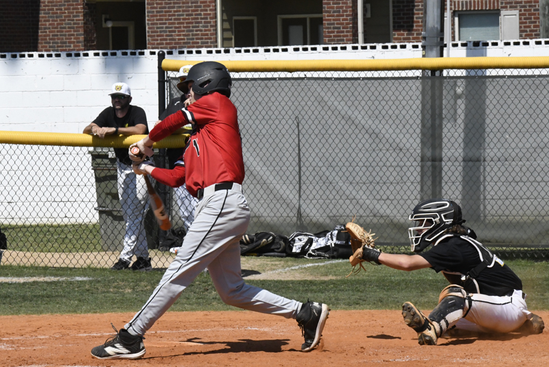 A baseball player in a red jersey swings at a pitch while the catcher crouches behind home plate during a game.