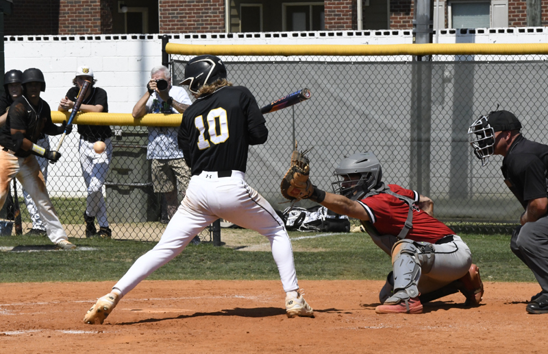 Baseball player in black jersey at bat, catcher in red gear ready to catch, umpire watching, and spectators behind the fence.