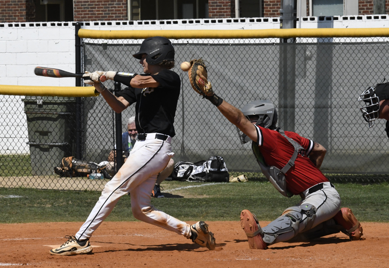 Baseball player in black jersey swings at a pitch as catcher in red gear reaches for the ball during a game.
